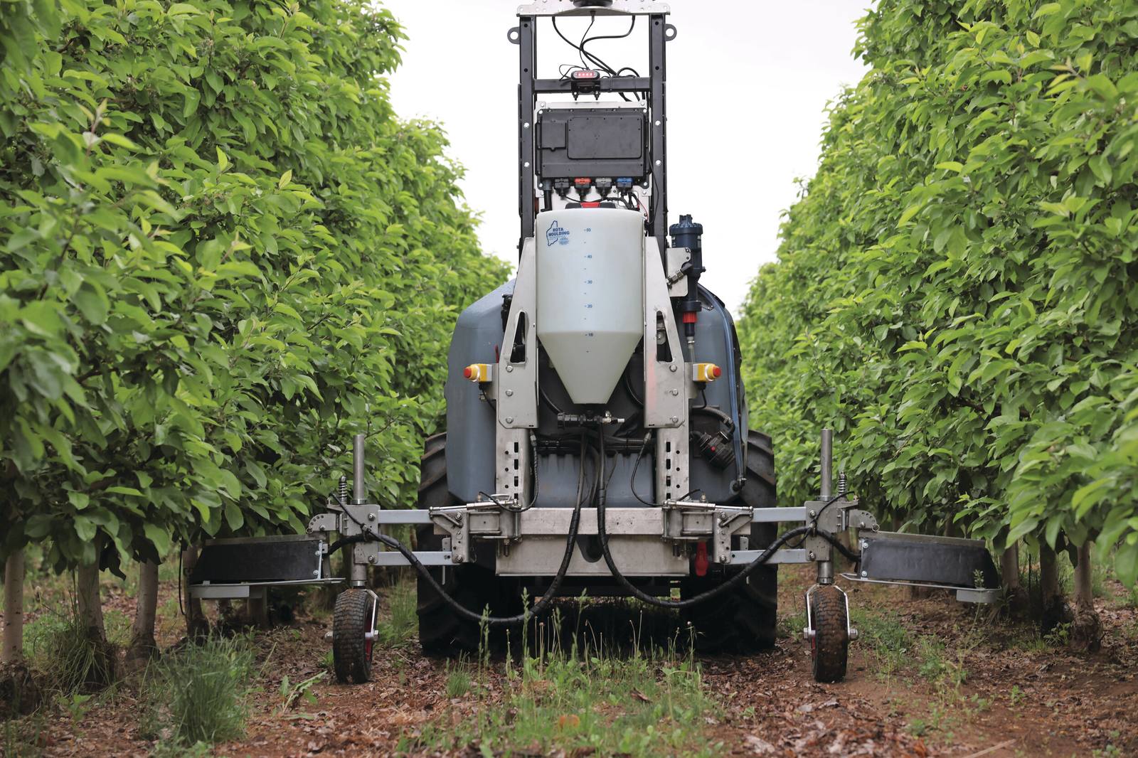 Yamaha Agriculture's Prospr autonomous hybrid vehicle fitted with the new Croplands Equipment herbicide attachment, with spray tank and breakaway boom arms, working an orchard row. The system automates nozzle activation and spray rates based on vehicle speed.